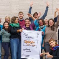 12 Personen stehen um das Plakat "Skills4Transformation" und winken fröhlich mit den Händen
